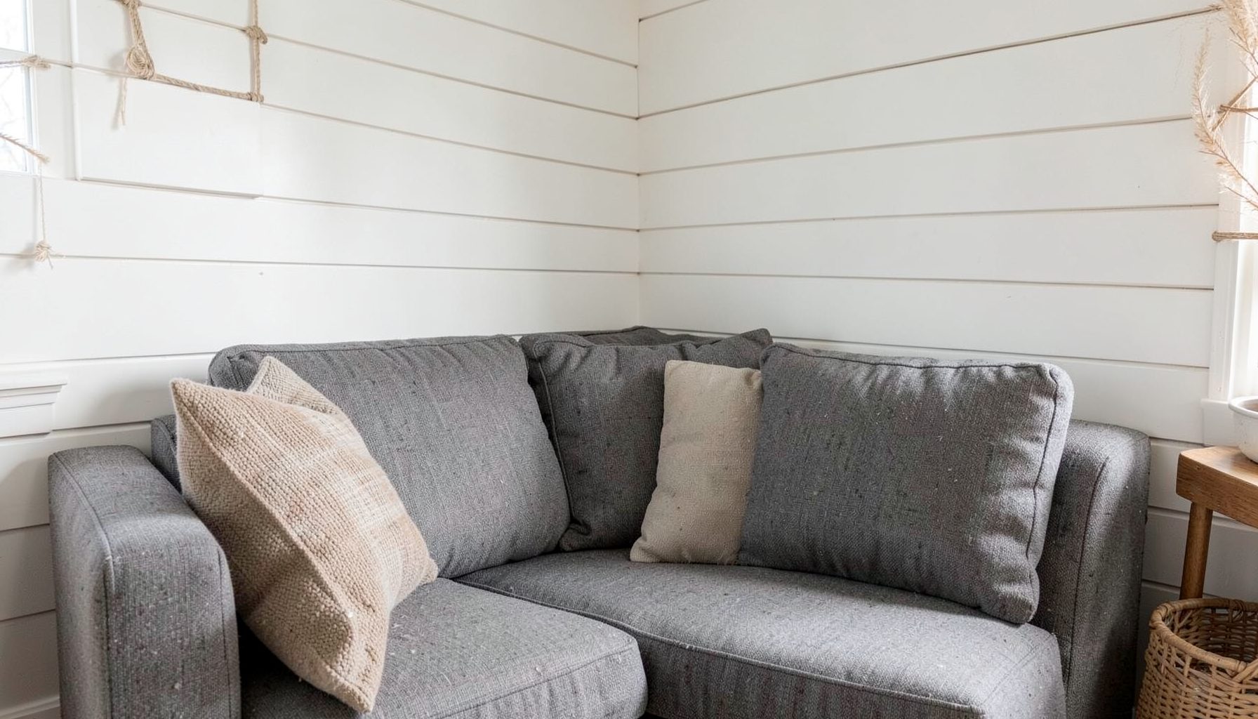 Cozy living room corner with a gray fabric sofa, white shiplap wall, and woven rattan accents in soft natural light