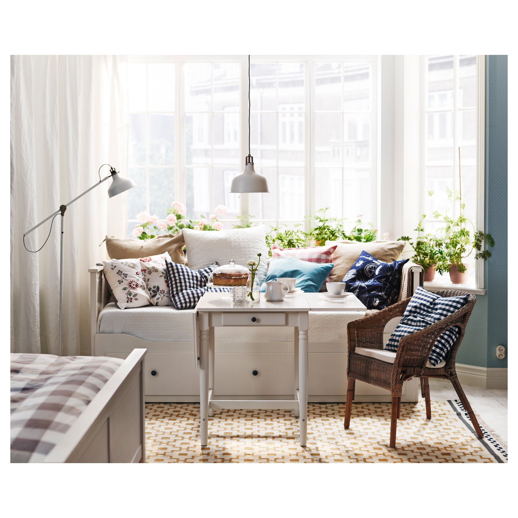 Bright sunroom with white daybed piled with patterned cushions, small drop-leaf table set for coffee, wicker chair