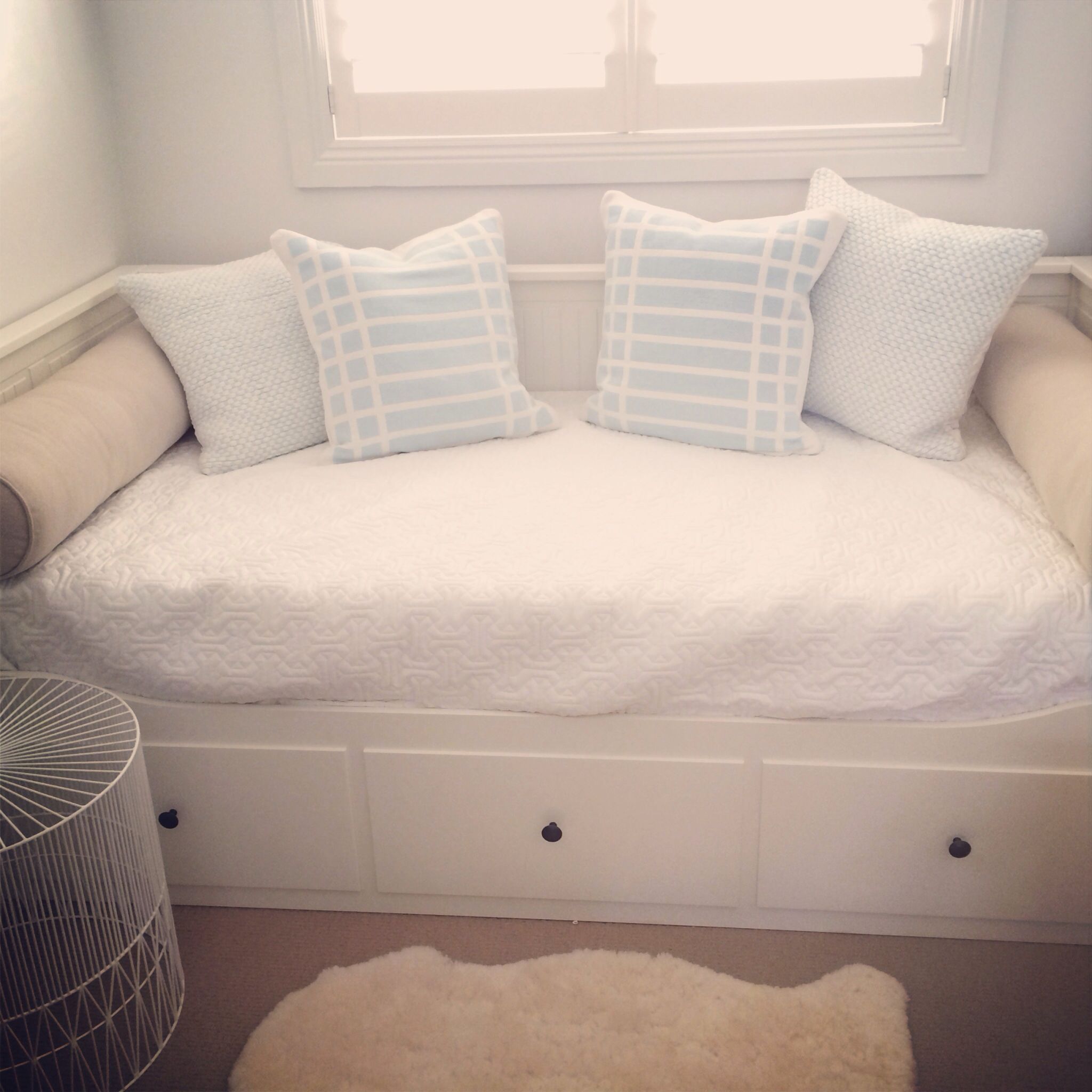 Simple bedroom nook with a white daybed featuring storage drawers, light blue and white patterned pillows, and a sheepskin rug