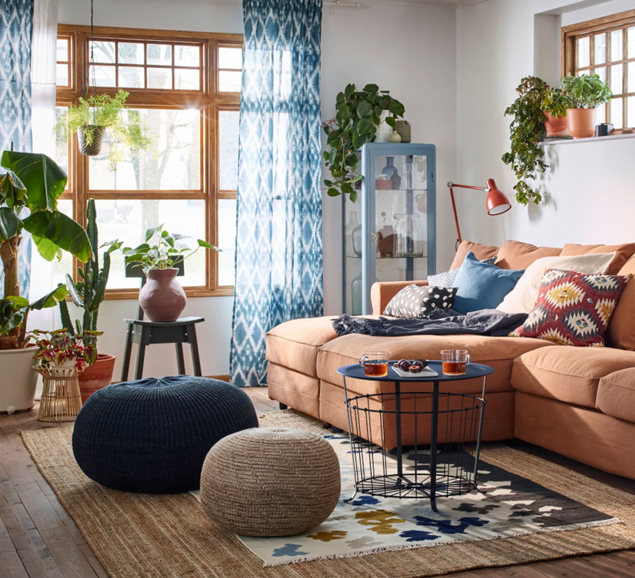 Living room with a rust-orange sectional sofa covered in patterned pillows, knit poufs, and a round wire side table