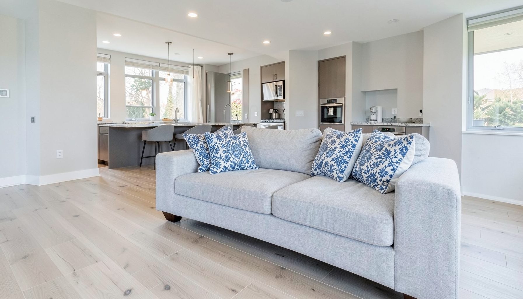 Open-plan living space featuring a light gray loveseat with blue and white patterned throw pillows against pale wood flooring