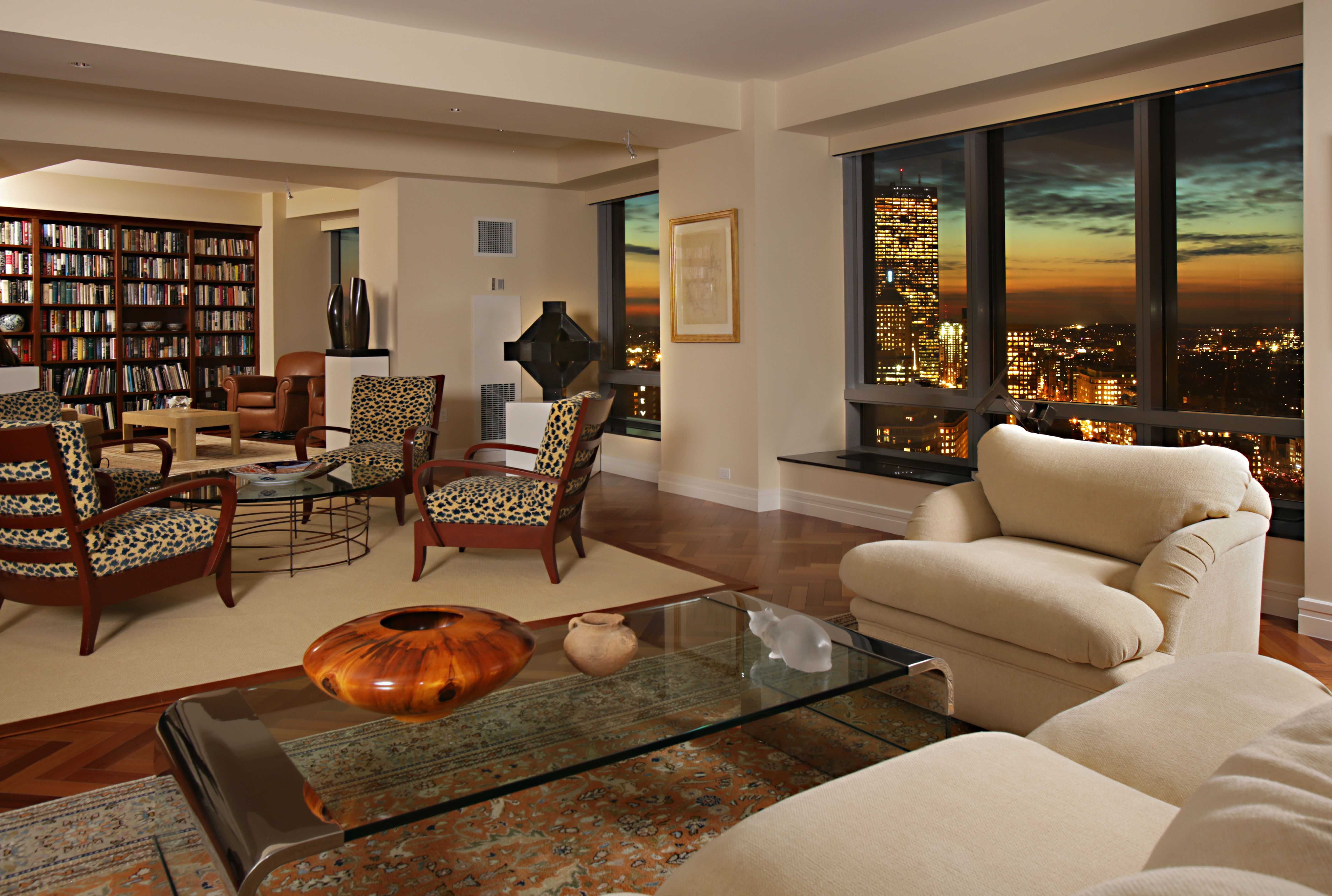 Upscale living room with cream armchairs, leopard-print chairs, bookshelves, and city skyline view at dusk