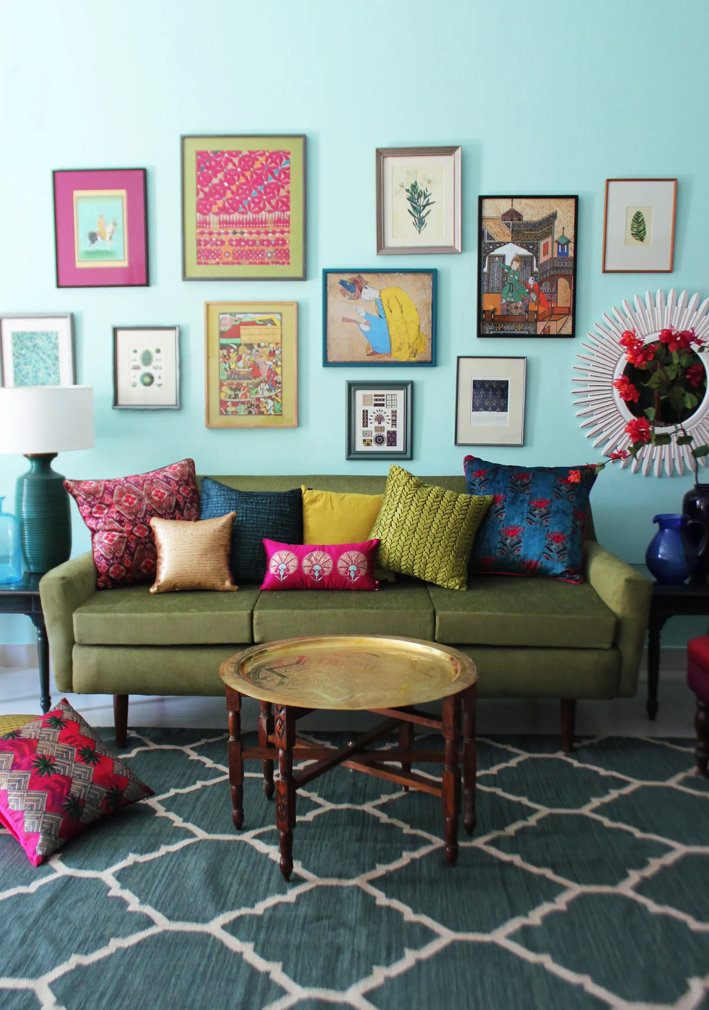 Bright turquoise living room wall covered in a gallery of colorful framed art, green sofa with jewel-toned patterned cushions